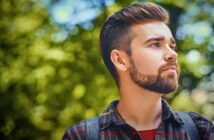 A bearded man with styled hair and a red plaid shirt looks into the distance against a blurred, leafy green background. He appears contemplative and outdoors in natural light.