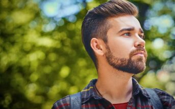 A bearded man with styled hair and a red plaid shirt looks into the distance against a blurred, leafy green background. He appears contemplative and outdoors in natural light.