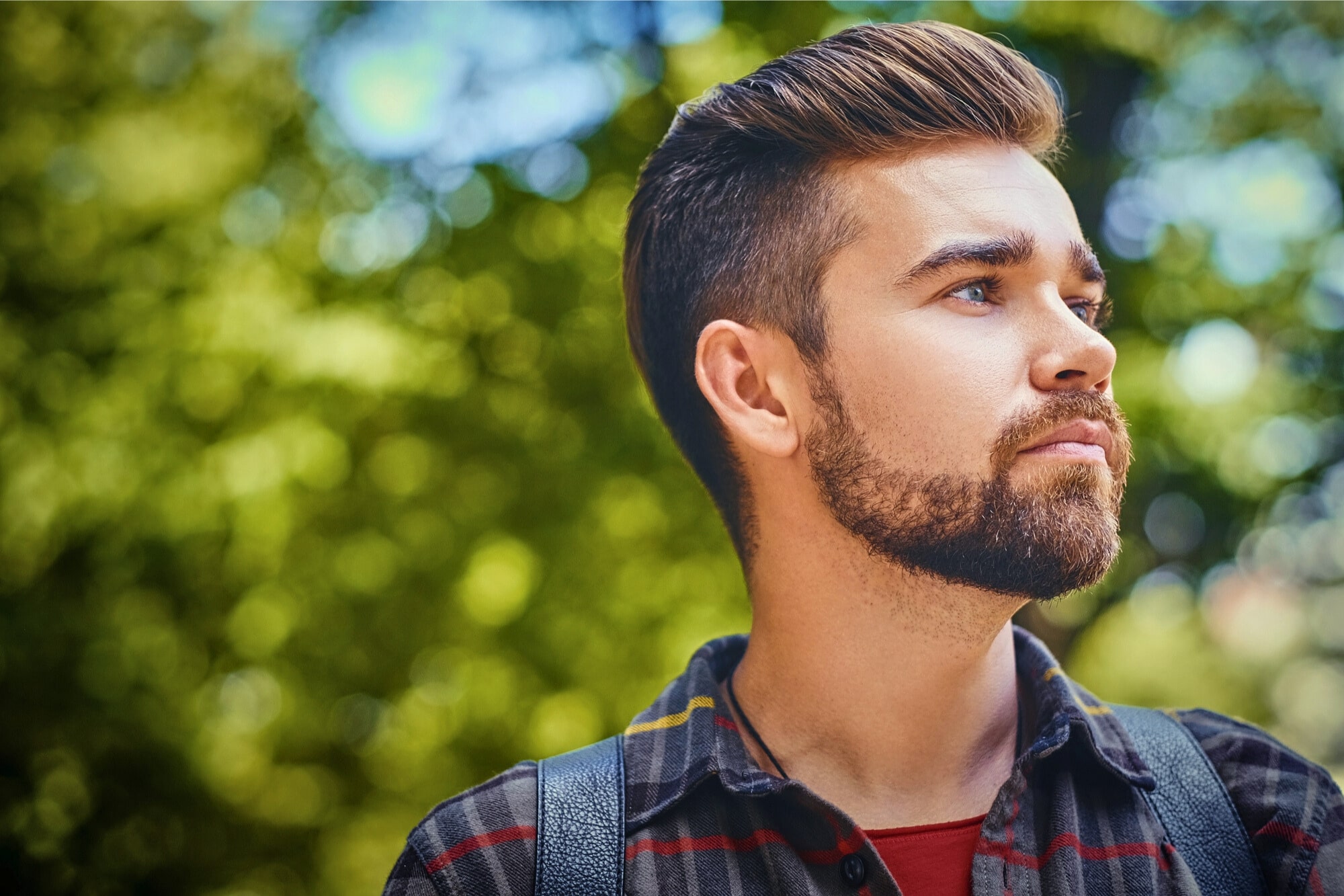 A bearded man with styled hair and a red plaid shirt looks into the distance against a blurred, leafy green background. He appears contemplative and outdoors in natural light.