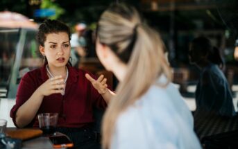 Two women are sitting at a table in a cafe. One with a red shirt gestures while speaking, appearing engaged in conversation. The other, facing away, listens attentively. A glass and a few items are on the table, with blurred outdoor scenery in the background.