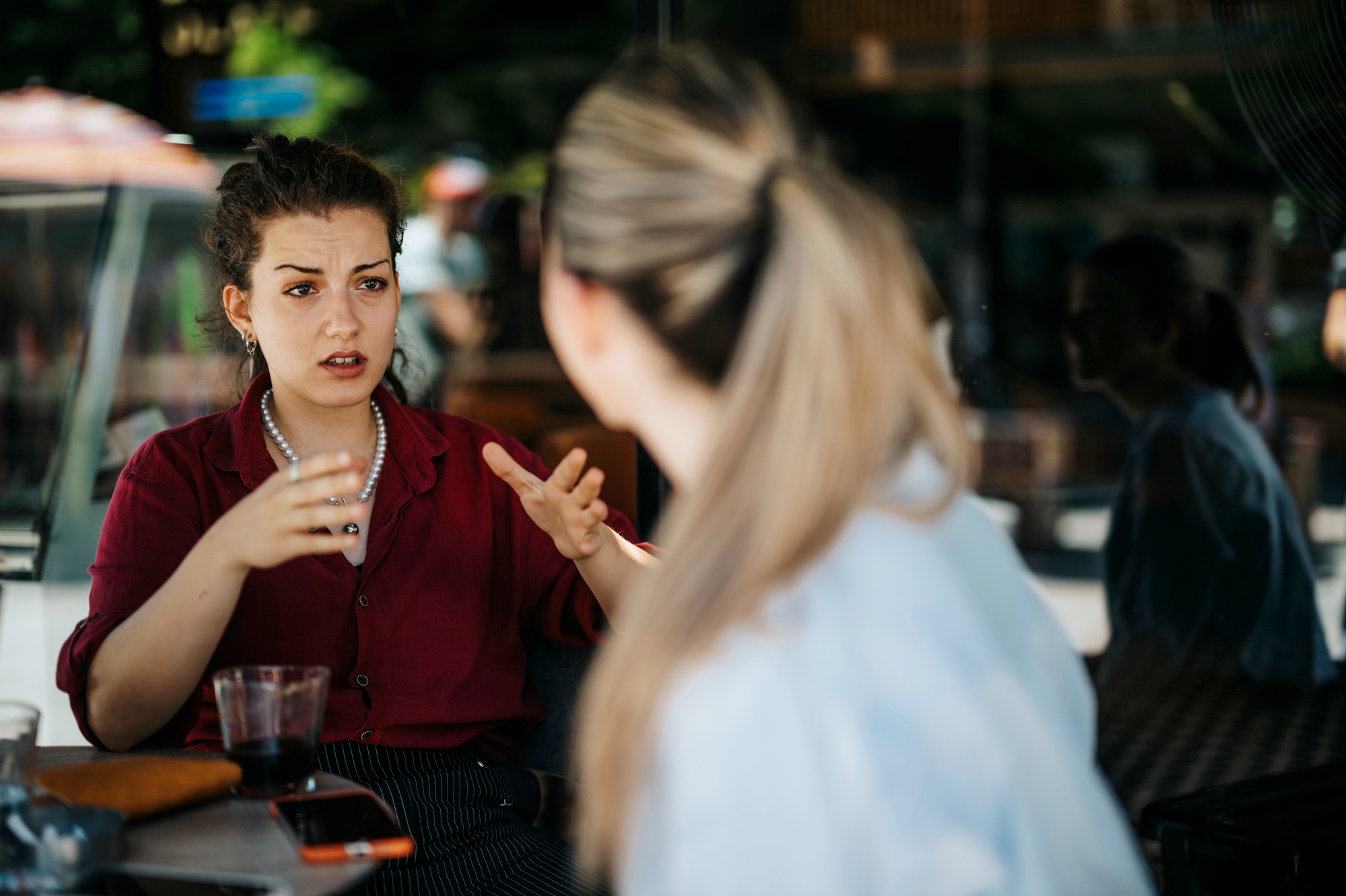 Two women are sitting at a table in a cafe. One with a red shirt gestures while speaking, appearing engaged in conversation. The other, facing away, listens attentively. A glass and a few items are on the table, with blurred outdoor scenery in the background.