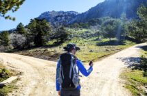 A person in hiking gear stands at a fork in a dirt trail, holding a GPS device. Surrounded by trees, the scene shows a mountainous landscape under a clear blue sky with the sun shining brightly.