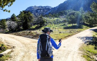 A person in hiking gear stands at a fork in a dirt trail, holding a GPS device. Surrounded by trees, the scene shows a mountainous landscape under a clear blue sky with the sun shining brightly.