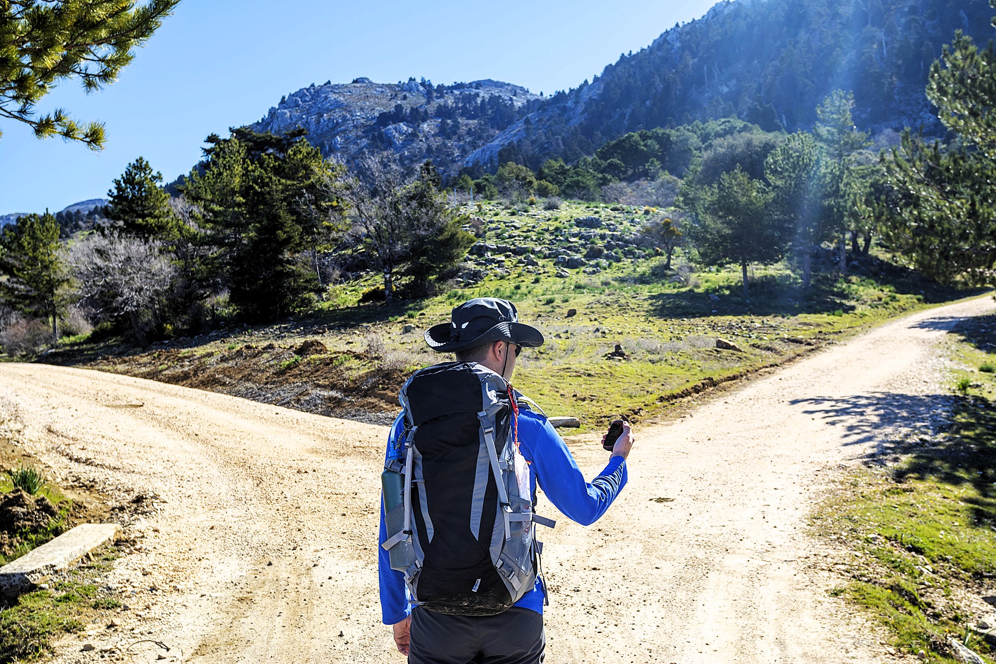 A person in hiking gear stands at a fork in a dirt trail, holding a GPS device. Surrounded by trees, the scene shows a mountainous landscape under a clear blue sky with the sun shining brightly.