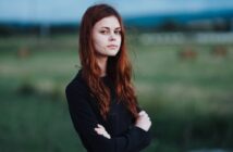 A person with long red hair stands outdoors, arms crossed, wearing a black top. The background features a blurred green field and a cloudy sky, conveying a serene and introspective atmosphere.