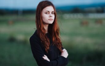 A person with long red hair stands outdoors, arms crossed, wearing a black top. The background features a blurred green field and a cloudy sky, conveying a serene and introspective atmosphere.