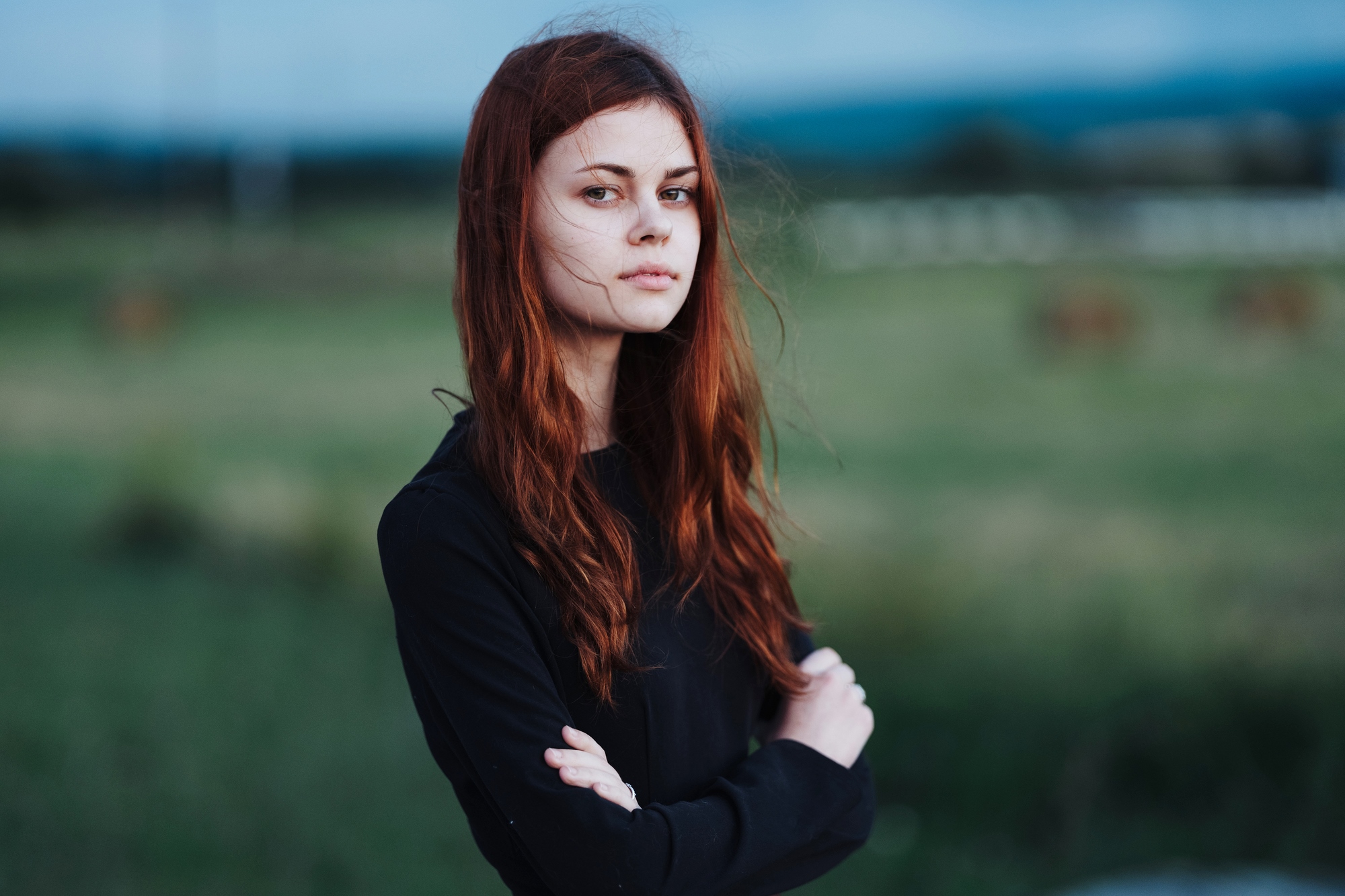 A person with long red hair stands outdoors, arms crossed, wearing a black top. The background features a blurred green field and a cloudy sky, conveying a serene and introspective atmosphere.