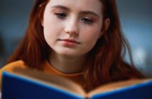 A person with long red hair and a thoughtful expression is reading a blue book. They are wearing a mustard yellow top, and the background is softly blurred.