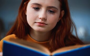 A person with long red hair and a thoughtful expression is reading a blue book. They are wearing a mustard yellow top, and the background is softly blurred.