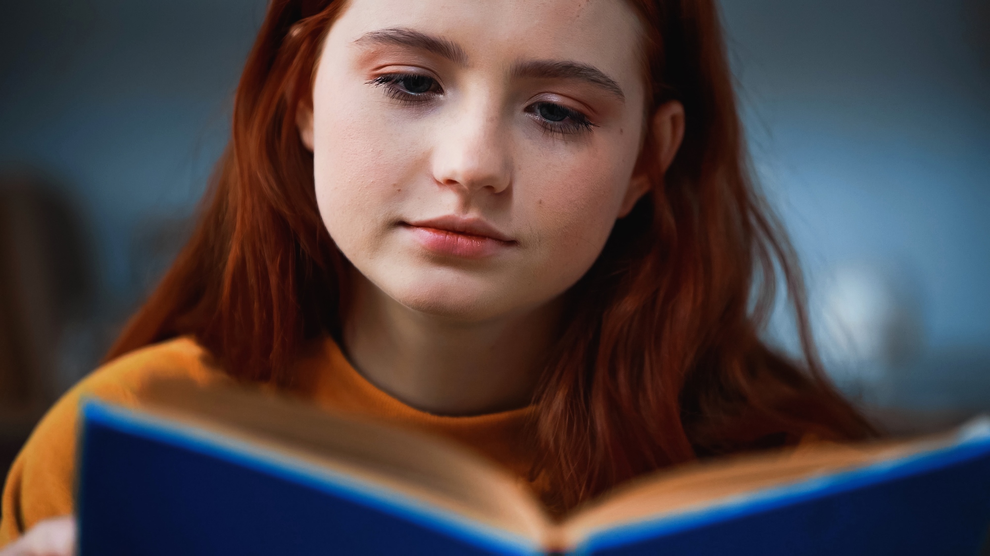 A person with long red hair and a thoughtful expression is reading a blue book. They are wearing a mustard yellow top, and the background is softly blurred.