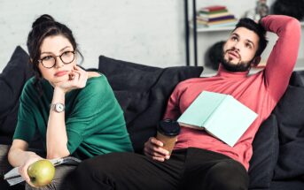 A woman and a man are sitting on a couch. The woman is wearing glasses, holding an apple, and looking away thoughtfully. The man is holding a coffee cup, with a book resting on his chest, looking upwards. Books are stacked in the background.