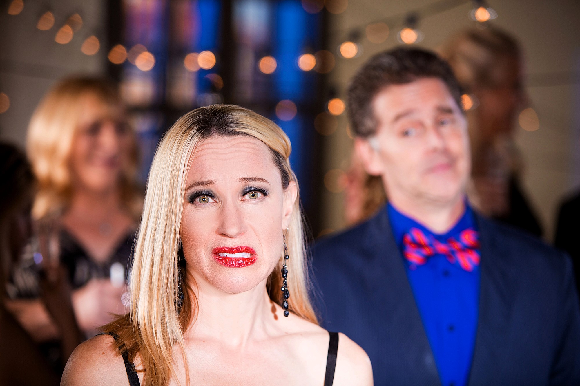 A woman with long blonde hair makes an awkward expression while standing at a party, adorned with string lights. A man in a blue shirt and patterned bow tie stands behind her, appearing unfocused and slightly perplexed. Other guests are blurred in the background.