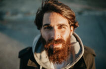 A bearded man with long hair and a warm smile is looking directly at the camera. He is wearing a dark coat over a light-colored hoodie. The background is blurred, suggesting an outdoor setting.