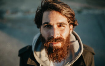 A bearded man with long hair and a warm smile is looking directly at the camera. He is wearing a dark coat over a light-colored hoodie. The background is blurred, suggesting an outdoor setting.