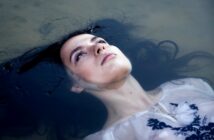 A woman in a white dress floats on water with her eyes open, looking contemplative. Her dark hair fans out around her, blending into the water. The sunlight softly illuminates her face.