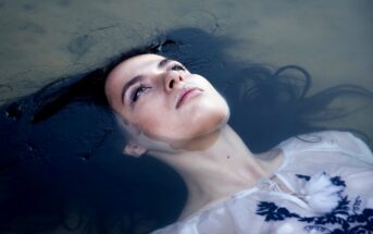 A woman in a white dress floats on water with her eyes open, looking contemplative. Her dark hair fans out around her, blending into the water. The sunlight softly illuminates her face.