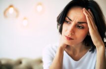 A woman with dark hair looks concerned, touching her forehead with one hand. She wears a white shirt. The background is softly lit with out-of-focus pendant lights.