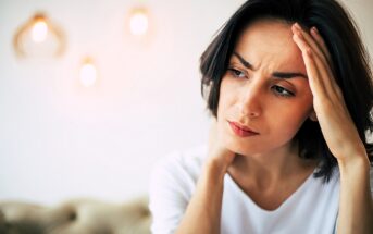 A woman with dark hair looks concerned, touching her forehead with one hand. She wears a white shirt. The background is softly lit with out-of-focus pendant lights.