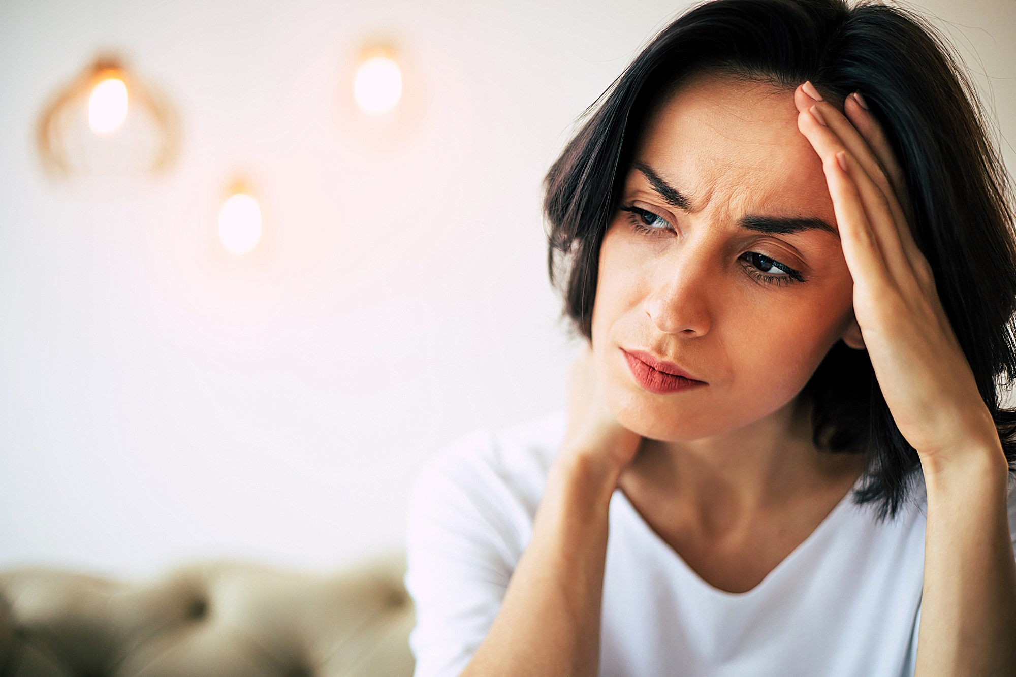 A woman with dark hair looks concerned, touching her forehead with one hand. She wears a white shirt. The background is softly lit with out-of-focus pendant lights.