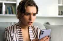 A woman sitting on a couch looks surprised while holding a smartphone. She is wearing a striped shirt, and there is a bookshelf in the background.