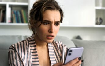 A woman sitting on a couch looks surprised while holding a smartphone. She is wearing a striped shirt, and there is a bookshelf in the background.