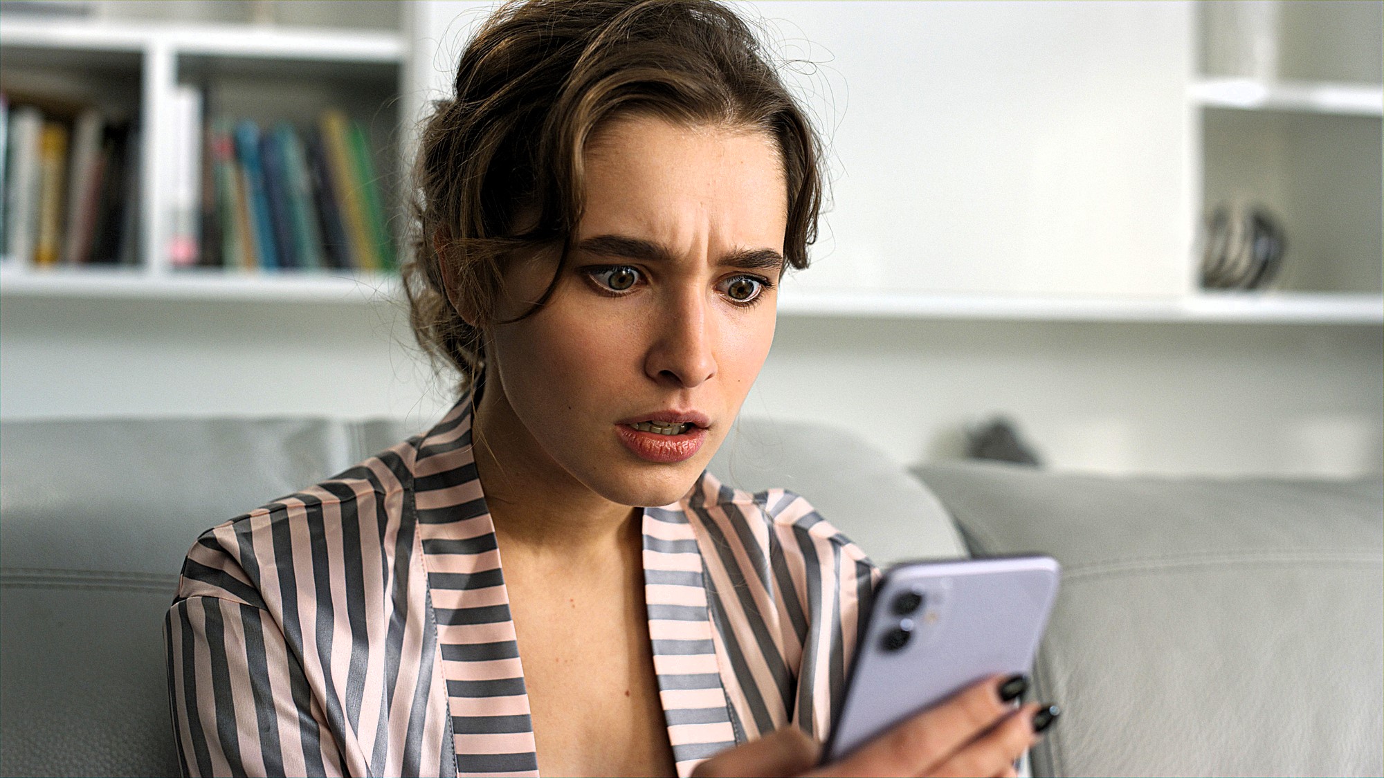 A woman sitting on a couch looks surprised while holding a smartphone. She is wearing a striped shirt, and there is a bookshelf in the background.