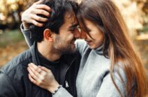 A couple embraces, smiling and touching foreheads. The man wears a black jacket, and the woman is in a gray sweater. They are outdoors, surrounded by blurred greenery and sunlight, creating a warm and intimate atmosphere.