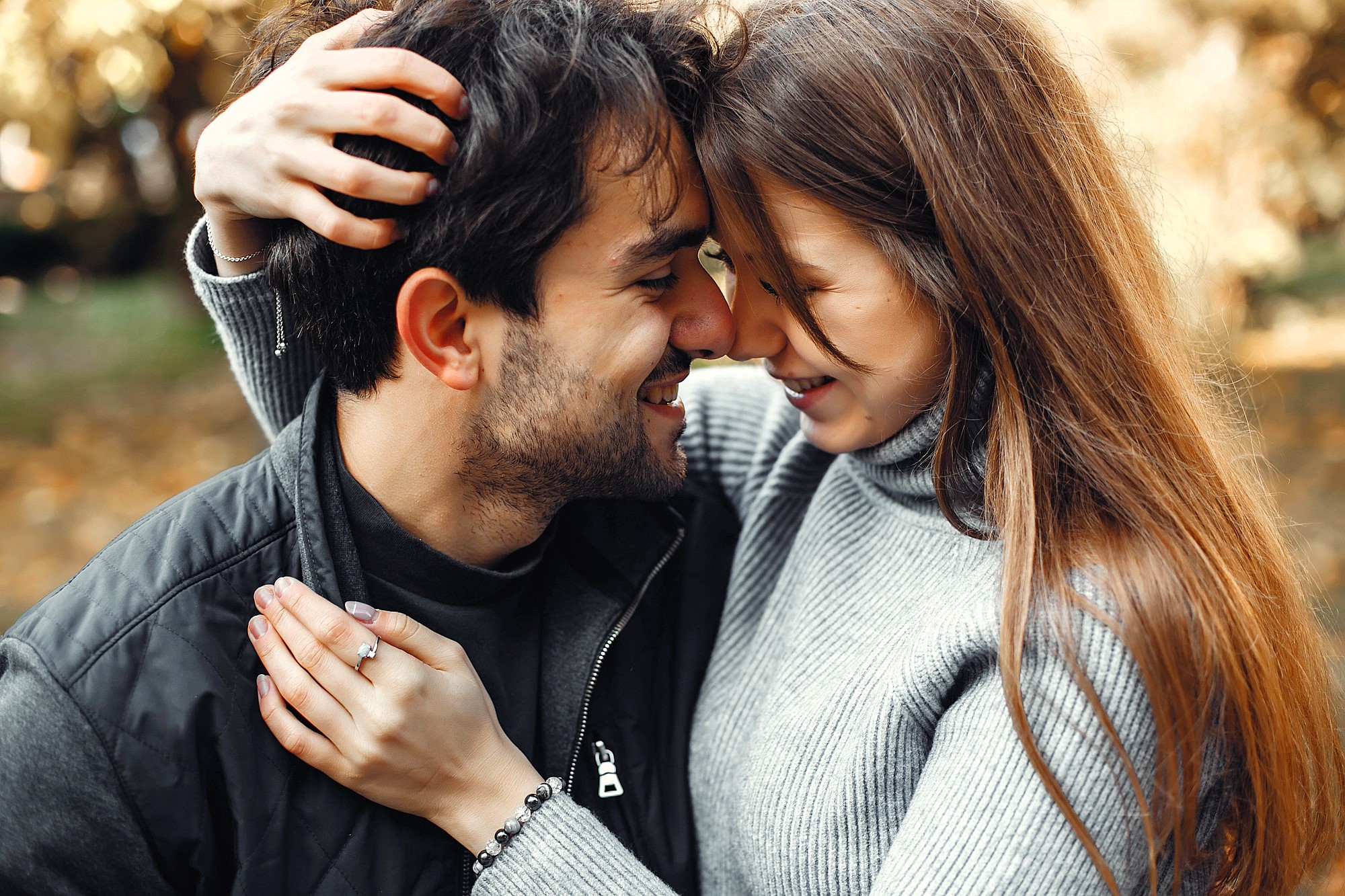 A couple embraces, smiling and touching foreheads. The man wears a black jacket, and the woman is in a gray sweater. They are outdoors, surrounded by blurred greenery and sunlight, creating a warm and intimate atmosphere.