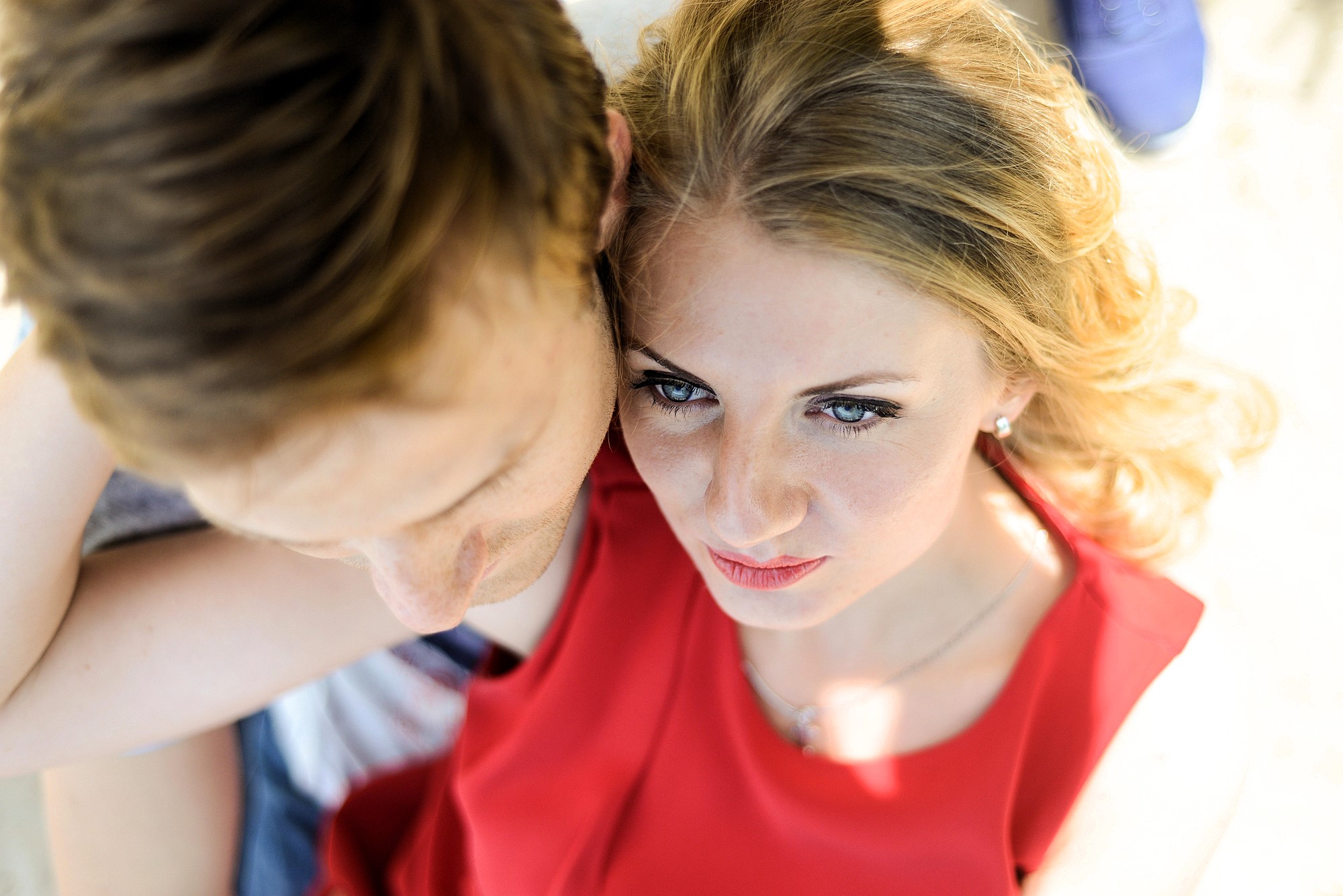 A woman in a red top and a man with light brown hair are sitting closely, viewed from above. The woman looks upward with a serious expression, while the man looks downward. Bright sunlight highlights their hair and faces.