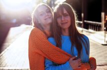 Two people smiling and embracing each other in the sunlight on a city street. One is wearing an orange sweater, the other a blue sweater. The sun creates a soft, glowing effect, and the background is slightly blurred.