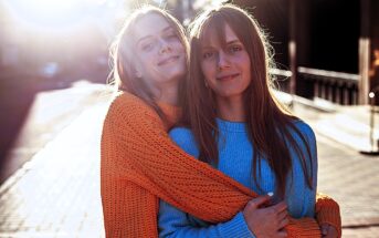 Two people smiling and embracing each other in the sunlight on a city street. One is wearing an orange sweater, the other a blue sweater. The sun creates a soft, glowing effect, and the background is slightly blurred.