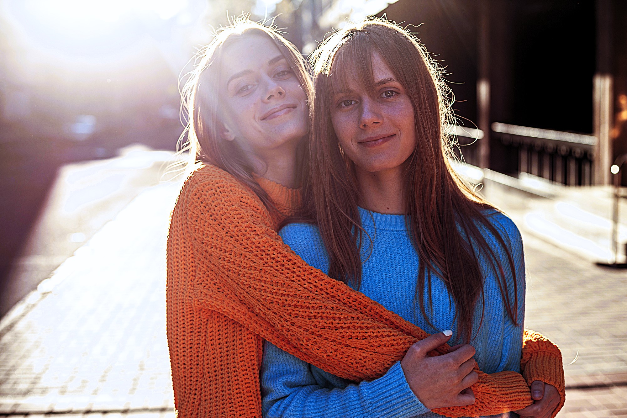 Two people smiling and embracing each other in the sunlight on a city street. One is wearing an orange sweater, the other a blue sweater. The sun creates a soft, glowing effect, and the background is slightly blurred.