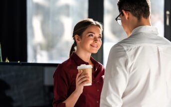 A woman in a maroon shirt holds a coffee cup and smiles at a man in glasses and a white shirt. They are indoors with natural light coming through large windows in the background.