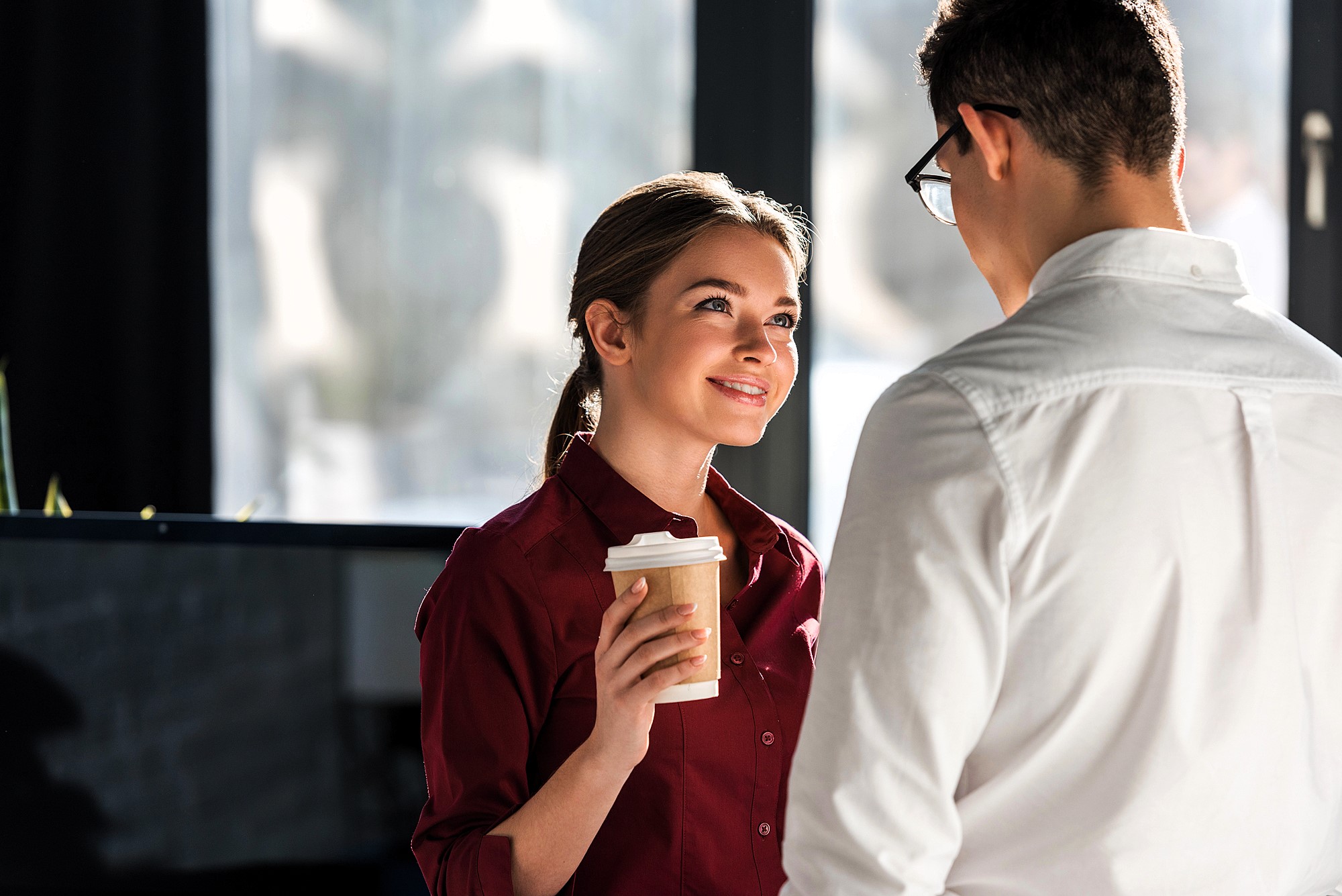 A woman in a maroon shirt holds a coffee cup and smiles at a man in glasses and a white shirt. They are indoors with natural light coming through large windows in the background.