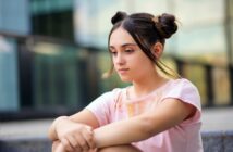 A young woman with dark hair in two buns sits on outdoor steps, wearing a pastel pink shirt. She appears thoughtful and relaxed, with a glass building in the blurred background.