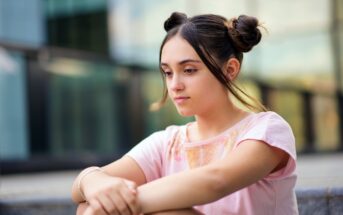 A young woman with dark hair in two buns sits on outdoor steps, wearing a pastel pink shirt. She appears thoughtful and relaxed, with a glass building in the blurred background.