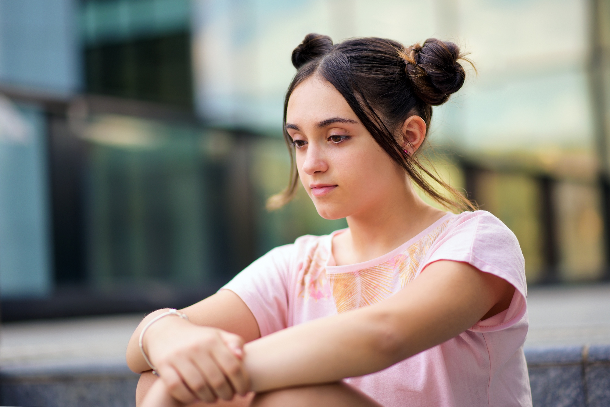 A young woman with dark hair in two buns sits on outdoor steps, wearing a pastel pink shirt. She appears thoughtful and relaxed, with a glass building in the blurred background.