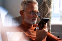 A man with gray hair and a beard, wearing glasses and a rust-colored sweater, sits in a chair and holds a phone to his mouth. Sunlight streams in through a window with patterned curtains in the background.