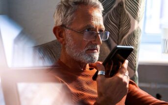 A man with gray hair and a beard, wearing glasses and a rust-colored sweater, sits in a chair and holds a phone to his mouth. Sunlight streams in through a window with patterned curtains in the background.