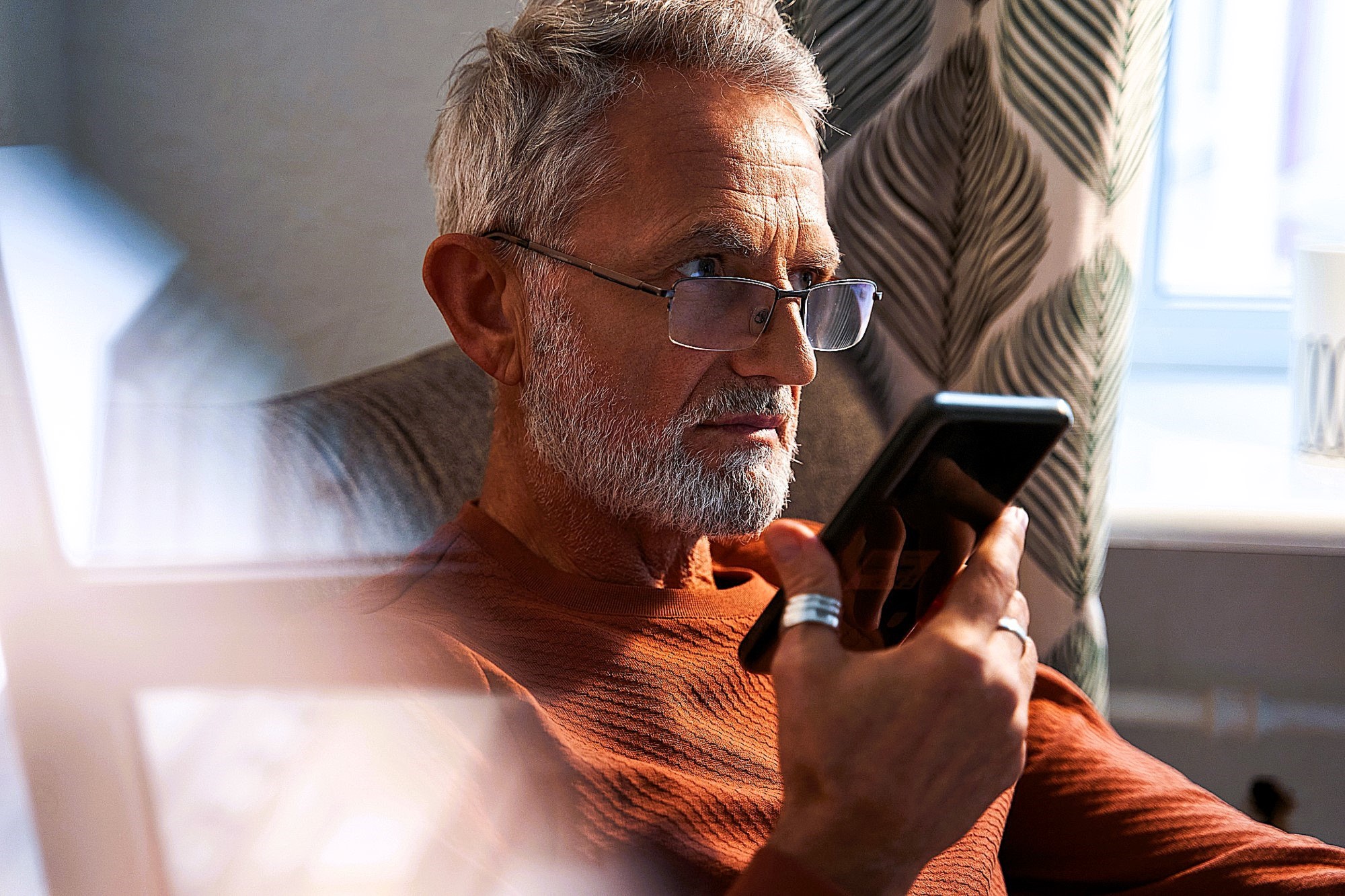 A man with gray hair and a beard, wearing glasses and a rust-colored sweater, sits in a chair and holds a phone to his mouth. Sunlight streams in through a window with patterned curtains in the background.