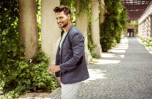 A man in a gray blazer and white pants smiles while walking through a sunlit outdoor corridor lined with greenery.