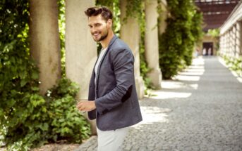 A man in a gray blazer and white pants smiles while walking through a sunlit outdoor corridor lined with greenery.