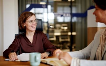 Two women in an office setting, one with glasses and a brown blouse, smiling while holding papers. The other, in a striped jacket, gestures toward an open notebook. A green mug sits on the wooden table. Blurred office background.