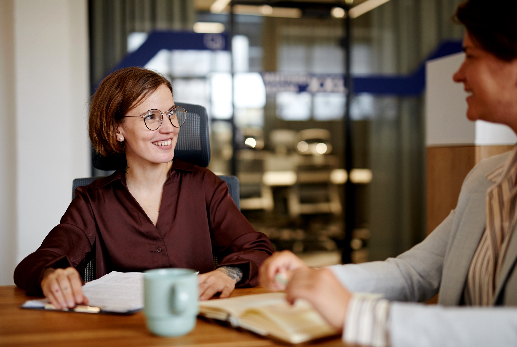 Two women in an office setting, one with glasses and a brown blouse, smiling while holding papers. The other, in a striped jacket, gestures toward an open notebook. A green mug sits on the wooden table. Blurred office background.