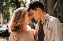 A couple stands close to each other, gently touching foreheads. They are outdoors on a sunny day, surrounded by trees and a blurred background. Both are smiling softly, with affectionate expressions.