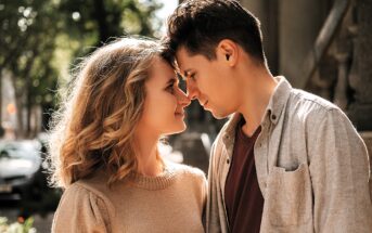 A couple stands close to each other, gently touching foreheads. They are outdoors on a sunny day, surrounded by trees and a blurred background. Both are smiling softly, with affectionate expressions.