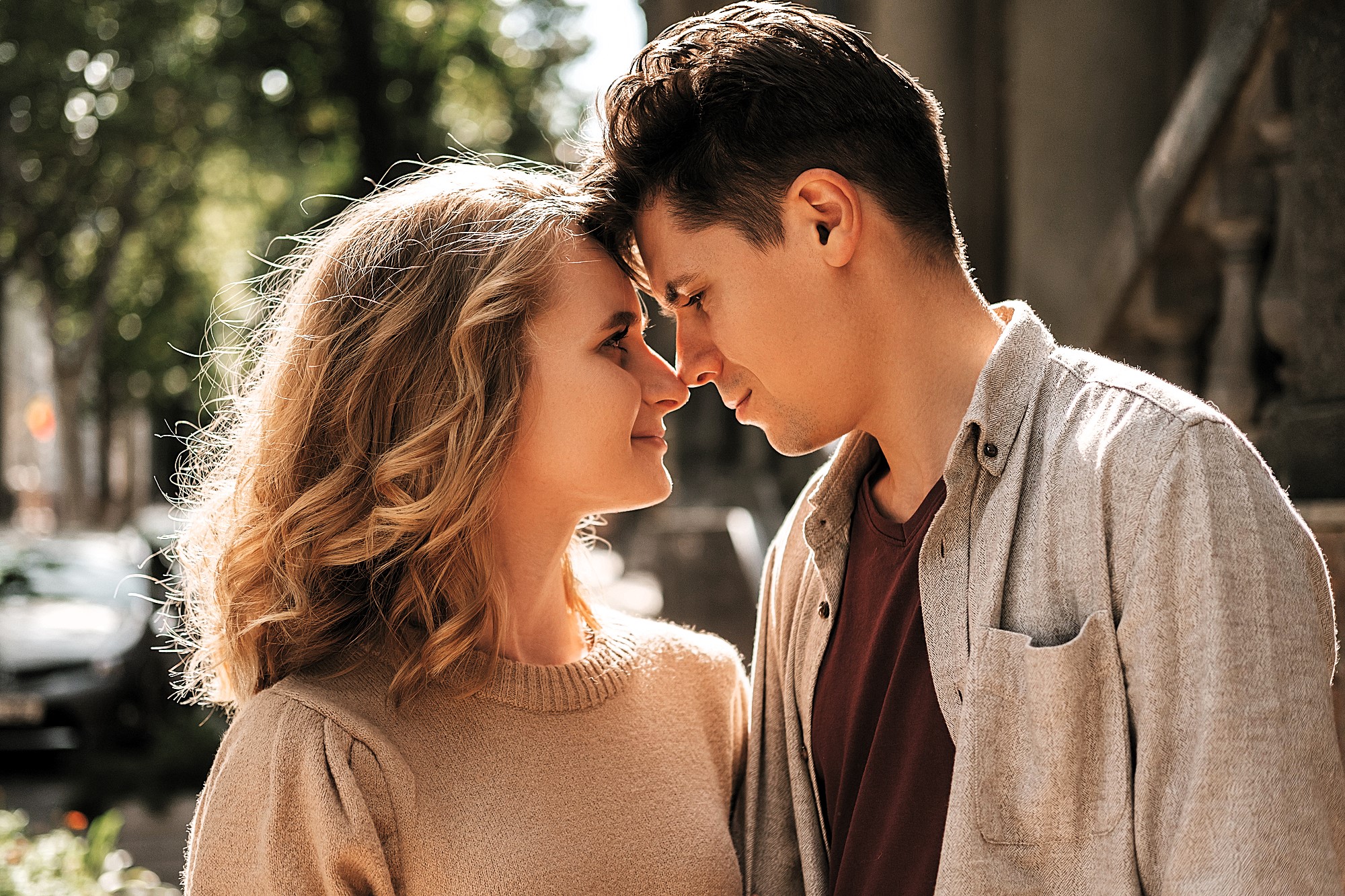 A couple stands close to each other, gently touching foreheads. They are outdoors on a sunny day, surrounded by trees and a blurred background. Both are smiling softly, with affectionate expressions.