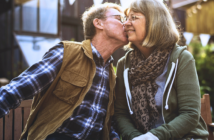 An elderly couple sits closely on a bench outdoors. The man, wearing a plaid shirt and brown vest, kisses the woman on the cheek. She wears glasses, a green jacket, and a scarf, smiling warmly. Sunlight and triangular banners are in the background.