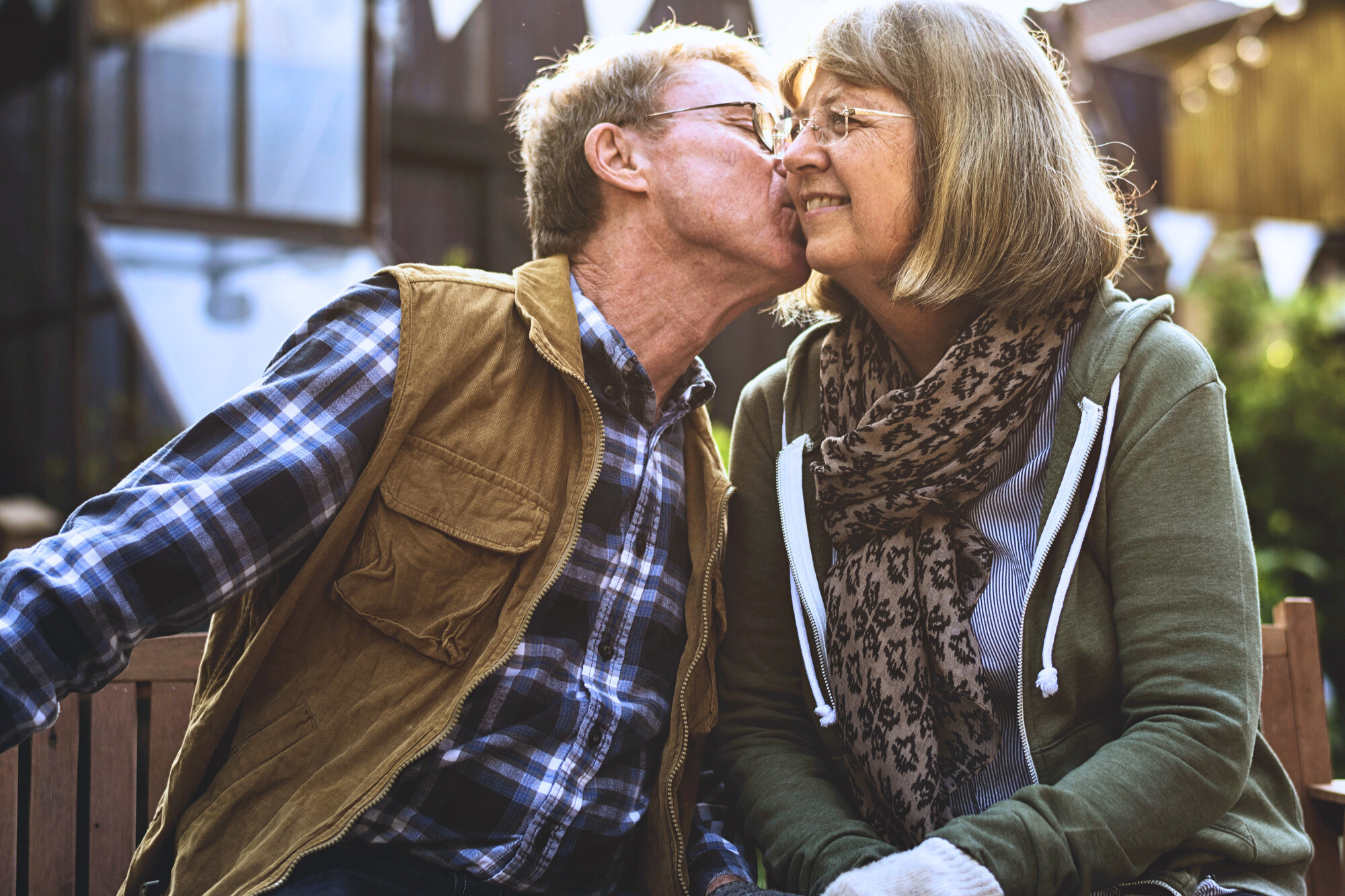 An elderly couple sits closely on a bench outdoors. The man, wearing a plaid shirt and brown vest, kisses the woman on the cheek. She wears glasses, a green jacket, and a scarf, smiling warmly. Sunlight and triangular banners are in the background.