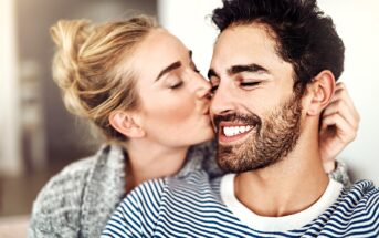 A woman with blonde hair is gently kissing a smiling, bearded man on the cheek. They are indoors, and the man is wearing a striped shirt while the woman wears a cozy sweater. The scene conveys warmth and affection.